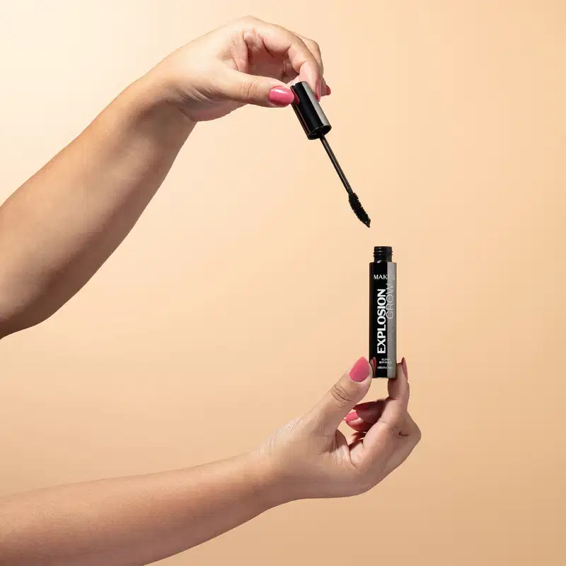 Uma mão feminina segura uma tampa com haste a aplicador na cor preta e a outra mão segura um tubo preto com letras brancas de mascara para cílios Make B. Explosion Grow em fundo em tons de rosa.