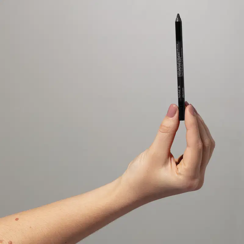 Mão feminina com unhas pintadas de lilás segurando um lápis para olhos Ultrablack Make B. em um fundo cinza.