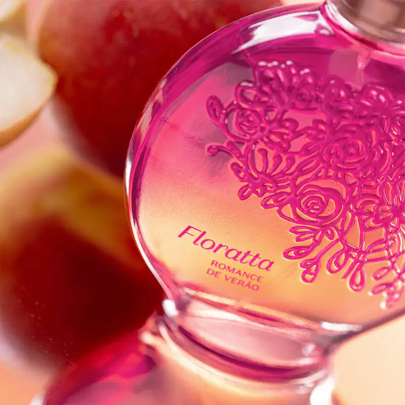 Vidro redondo com líquido rosa, floral em relevo rosa escuro, pulverizador prateado e tampa transparente contendo desodorante colônia Floratta Romance de Verão sobre superfície desfocada em vermelho e amarelo.
