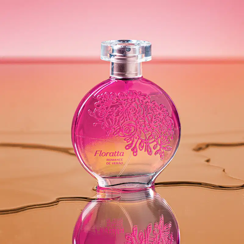Vidro redondo com líquido rosa, floral em relevo rosa escuro, pulverizador prateado e tampa transparente contendo desodorante colônia Floratta Romance de Verão refletido em superfície de vidro molhada.