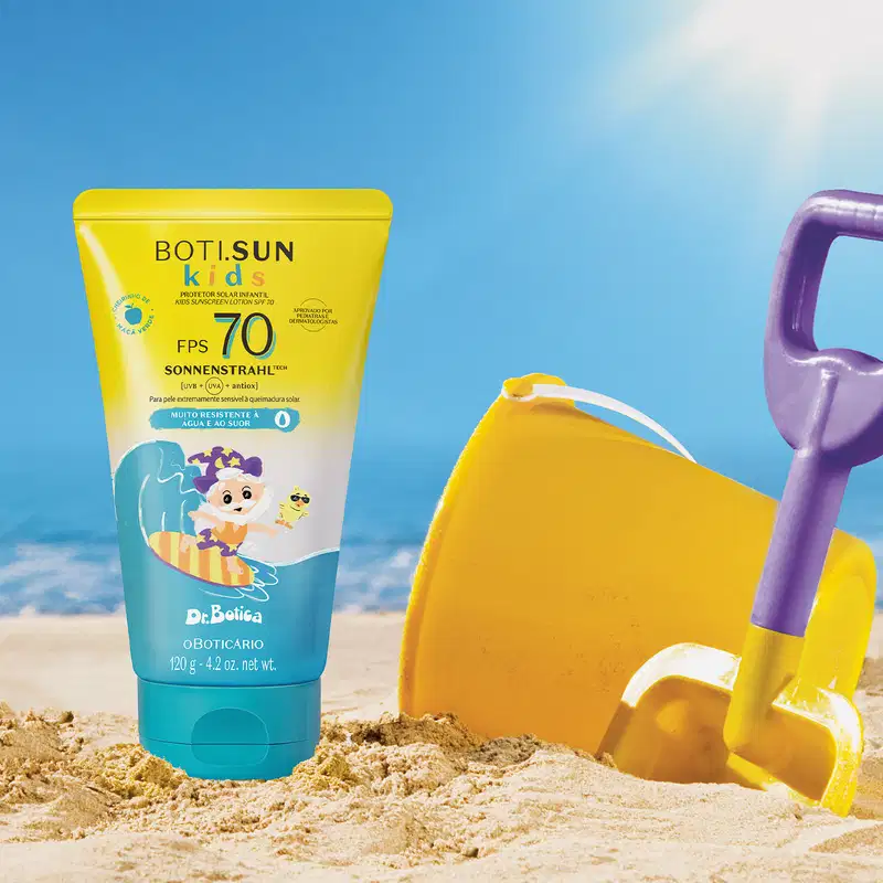Imagem de um tubo de protetor solar infantil Boti Sun Kids FPS 70 em praia com brinquedos de areia amarelos e roxos, sob céu azul e sol brilhante com o mar ao fundo.