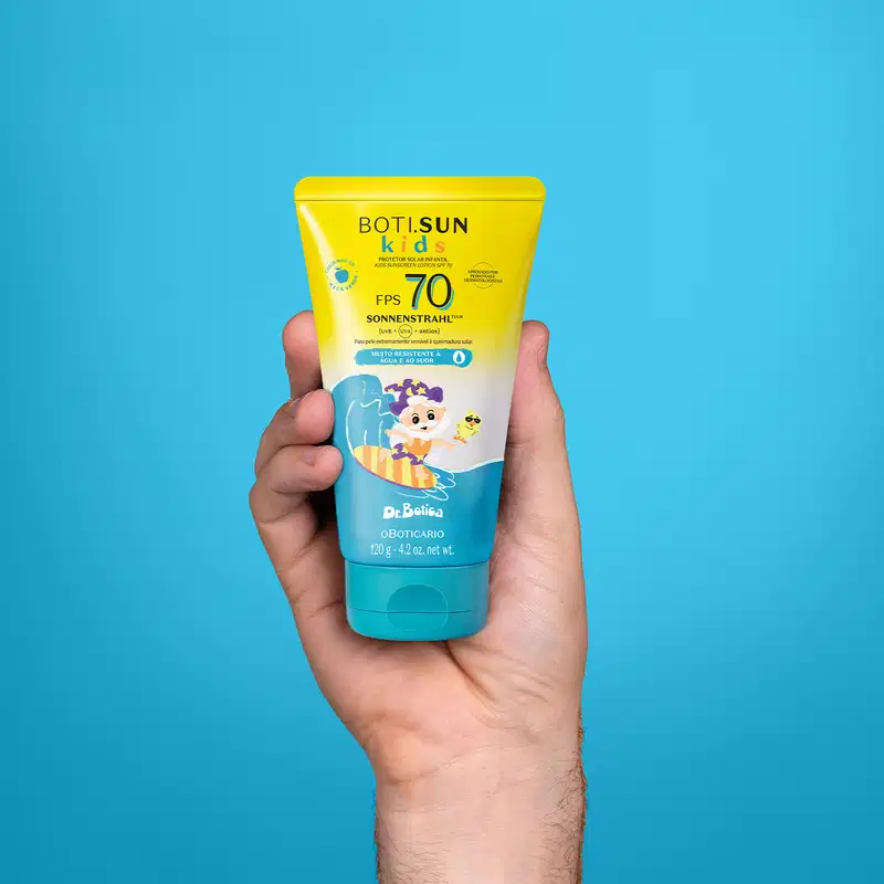 Imagem de uma mão segurando um frasco amarela e azul de protetor solar infantil Boti Sun Kids com FPS 70 com rótulo ilustrado de personagens da marca Dr. Botica em fundo azul.