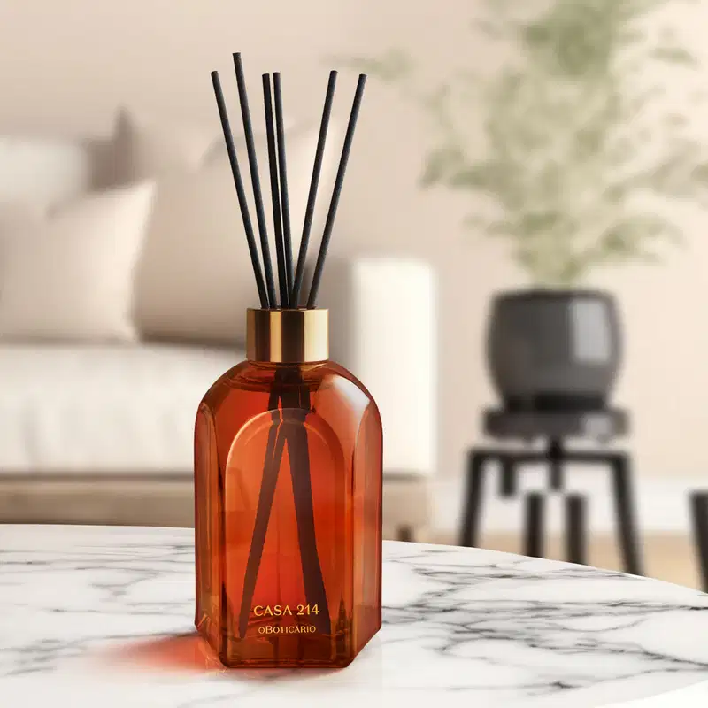 Difusor de ambientes em vidro laranja com varetas e letras douradas da marca Casa 214 Vanilla Sublime sobre superfície de mármore branco e cinza, com objeto preto desfocado ao fundo.