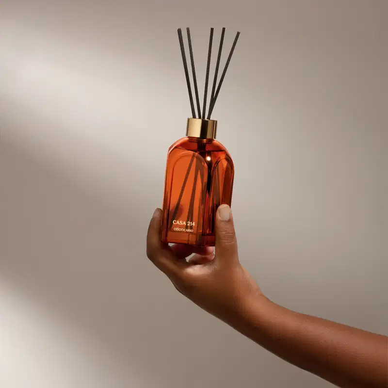 Mão segurando um Difusor de ambientes em vidro laranja com varetas e letras douradas da marca Casa 214 Vanilla Sublime em fundo cinza com sombras.
