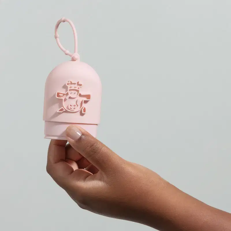 Mão segurando uma capinha de silicone rosa para creme de mãos da marca Cuide-se Bem Deleite, com ilustração de uma vaquinha em relevo e alça para pendurar. O fundo da imagem é azul claro.
