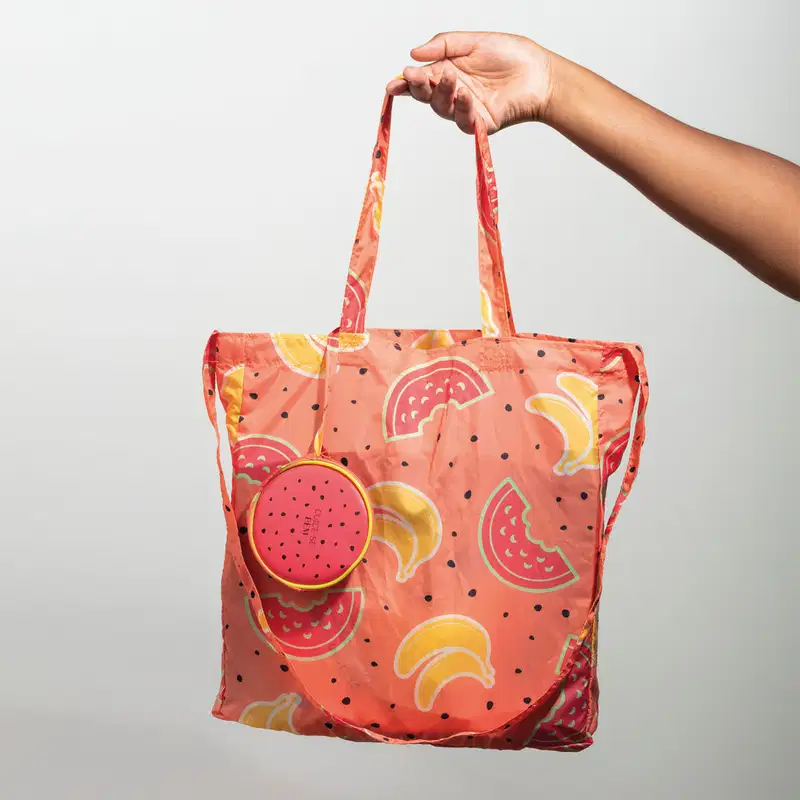 Mão segurando uma Sacola com alça de ombro e transversal na cor rosa com ilustrações de melancias, bananas e sementes pretas da marca Cuide-se Bem Feira em fundo cinza.
