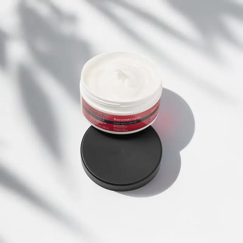 Pote branco aberto com creme branco dentro dele e tampa preta apoiada na embalagem. O produto possui rótulo na cor vermelha metalizada, preta e branca. A embalagem está em fundo claro com sombra de folhagens.