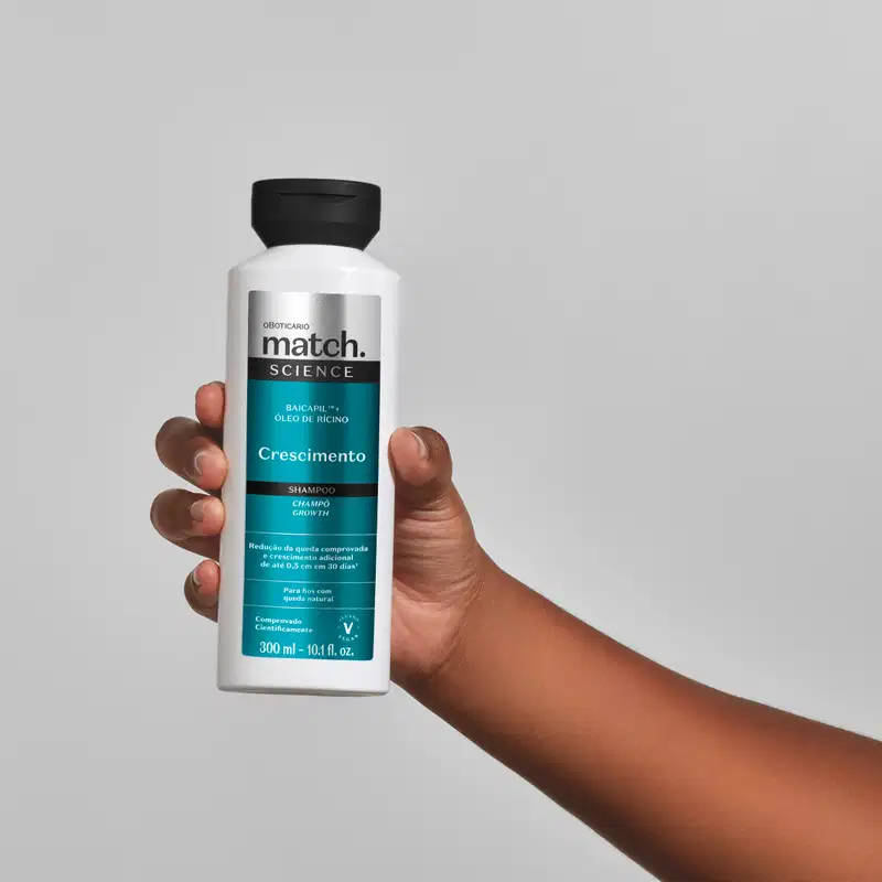 Mão segurando um vidro branco com tampa preta e rótulo azul, preto e prateado de shampoo Match Science Crescimento em fundo cinza.