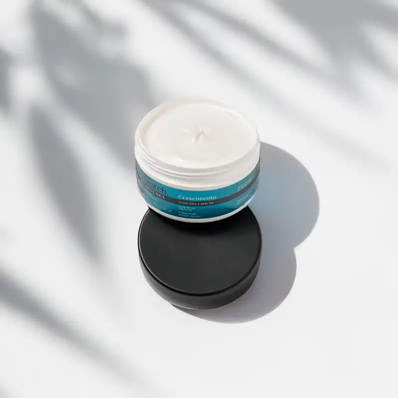 Pote branco aberto com creme branco dentro dele e tampa preta apoiada na embalagem. O produto possui rótulo na cor azul metalizado, preto e branco. A embalagem está em fundo claro com sombra de folhagens.