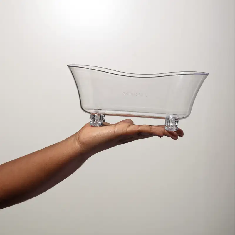 Mão feminina segurando uma banheira em acrílico branco em formato oval alongado com bordas arredondadas em fundo cinza claro.