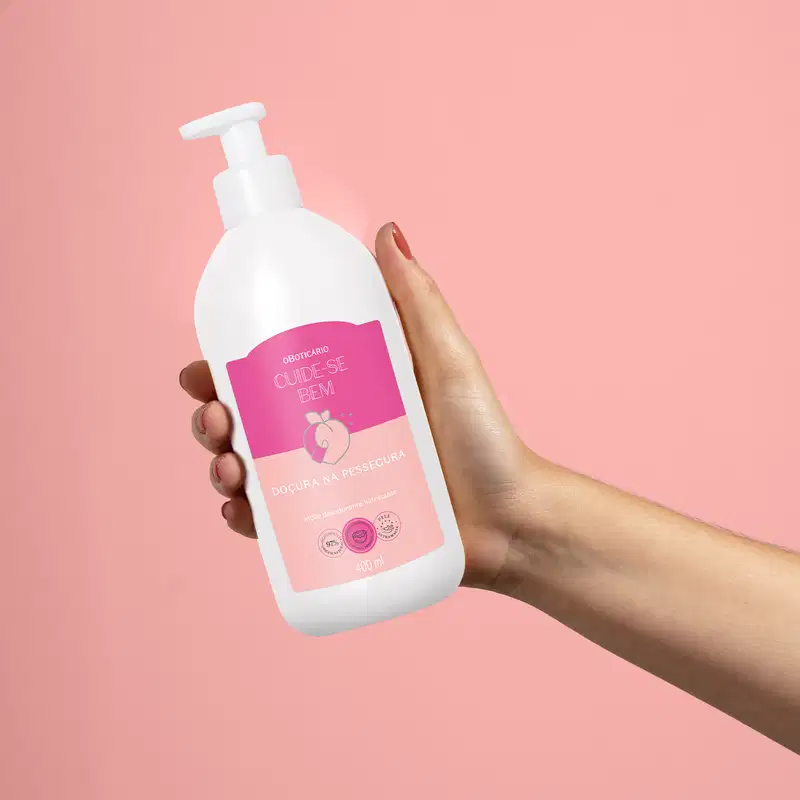 Frasco branco contendo um produto da linha "Cuide-se Bem" sendo segurado por mão feminina em fundo rosa. O rótulo do produto é predominantemente rosa, com o texto "Cuide-se Bem" e "Doçura na Pele Segura".