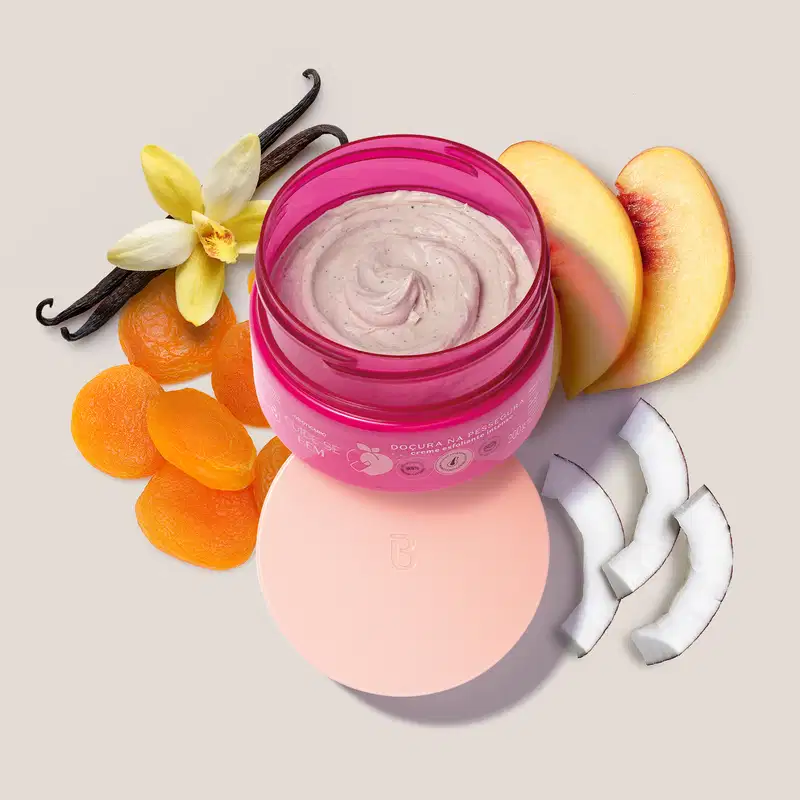 Pote pink creme esfoliante aberto com tampa rosa posicionada embaixo dele e diversos ingredientes naturais, como flores de baunilha, fatias de pêssego e rodelas de coco ao redor, sugerindo que o produto possui uma fórmula com ingredientes naturais.