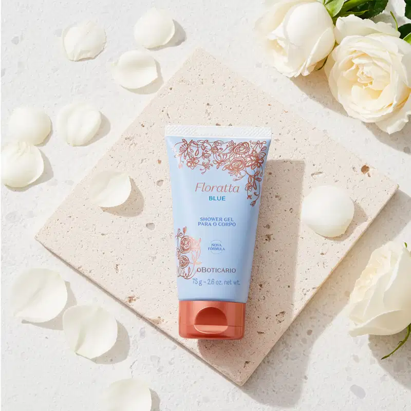 Imagem de um tubo de gel de banho da marca Floratta Blue sobre superfície de pedra clara, com pétalas de rosas brancas ao redor e buquê de rosas brancas ao fundo. O cenário sugere um produto delicado e feminino.