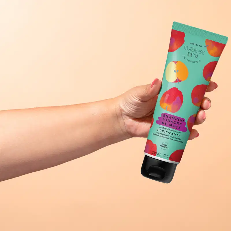 Imagem da embalagem do Shampoo Cuide-se Bem Feira Vinagre de Maçã em tons de verde, com ilustrações de maçãs, sendo segurado por mão em fundo bege claro.