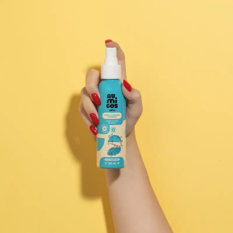 Mão segurando um Frasco em tons de azul e rosa com tampa spray branca e letras pretas contendo 120ml de limpa e hidrata patinhas da marca Au.Migos em fundo alaranjado.