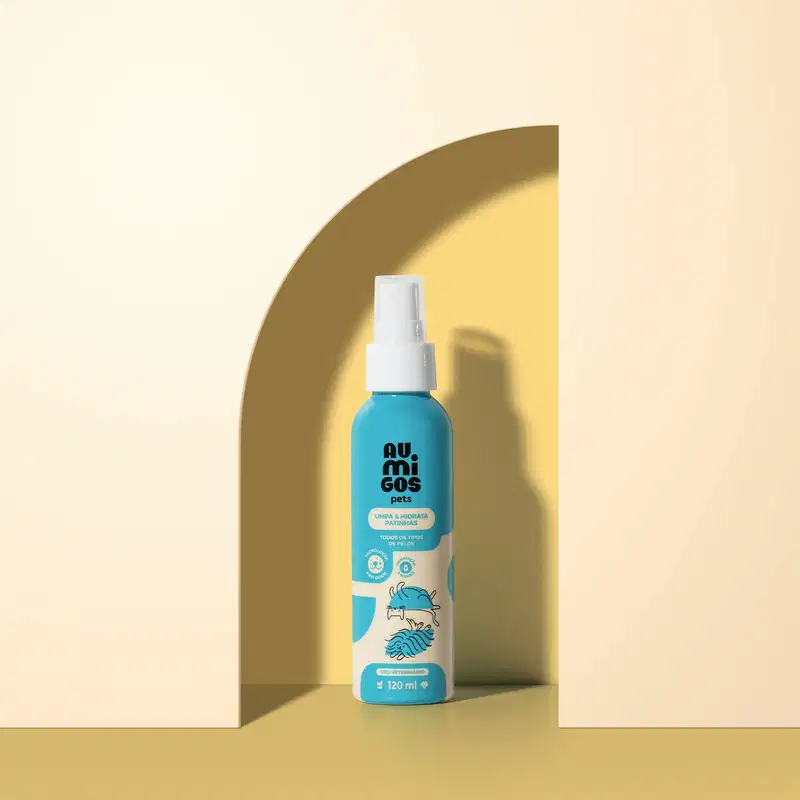 Frasco em tons de azul e rosa com tampa spray branca e letras pretas contendo 120ml de limpa e hidrata patinhas da marca Au.Migos. O produto está dentro de um arco com fundo e superfície em tons de laranja.