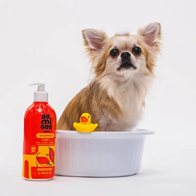Frasco laranja e rosa com ilustração de pets no rótulo e bomba dosasdora branca contendo 400ml de shampoo 5 em 1 da marca Au.Migos Pets. Ao lado, um cachorro está em uma banheira branca com um patinho de borracha perto dele.