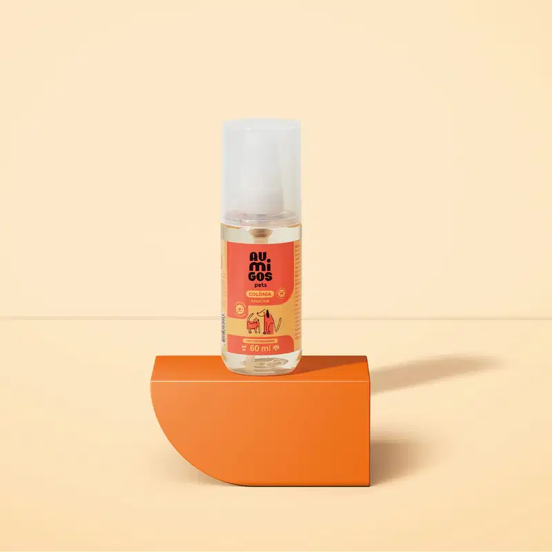 Frasco transparente com líquido claro, válvula spray branca, tampa transparente e rótulo laranja e rosa com ilustrações de um gato e de um cachorro contendo colônia adultos da marca Au.Migos Pets. O produto está em uma plataforma e fundo laranja.
