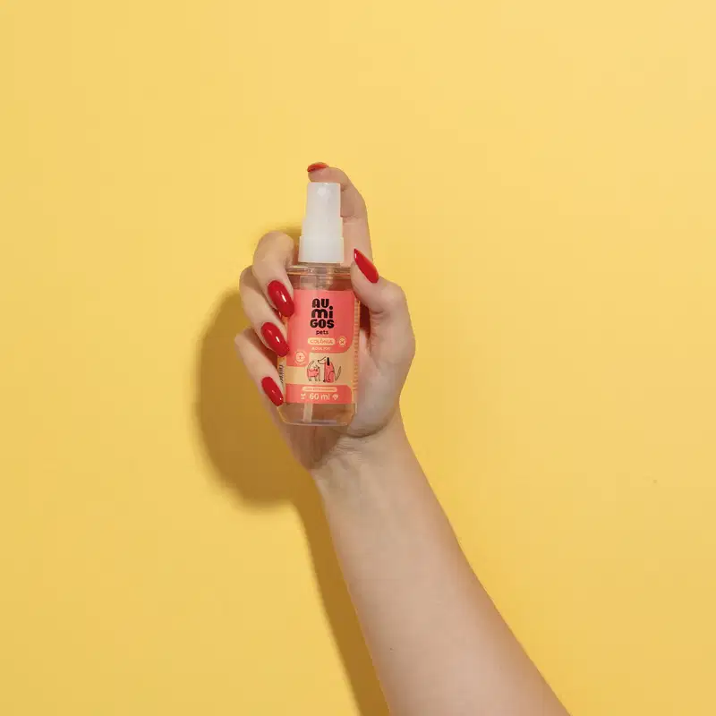 Mão segura um frasco com líquido claro, válvula spray branca e rótulo laranja e rosa com ilustrações de um gato e de um cachorro contendo 60ml de colônia adultos da marca Au.Migos Pets em fundo alaranjado.