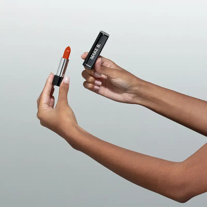Uma mão feminina está segurando um Batom glossy na cor Coral da marca Make B. em embalagem prata e preta e a outra mão segura a tampa na cor preta com letras prateadas. O fundo da imagem é cinza.