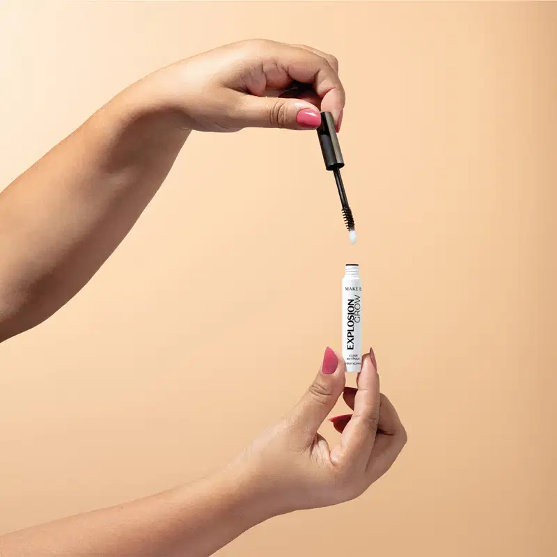 Duas mãos femininas, uma segurando uma tampa com aplicador na cor preta e a outra segura um tubo branco com letras pretas de sérum para cílios e sobrancelhas Explosion Grow da marca Make B em fundo em tons de rosa.