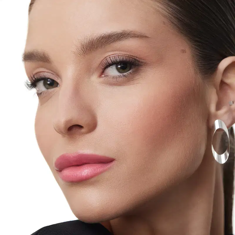 A imagem mostra um close-up do rosto de uma mulher usando maquiagem. Ela tem sobrancelhas bem delineadas em um tom castanho escuro. Seus olhos estão realçados com sombra marrom delineador e cílios volumosos. Os lábios estão com um batom rosado brilhante.