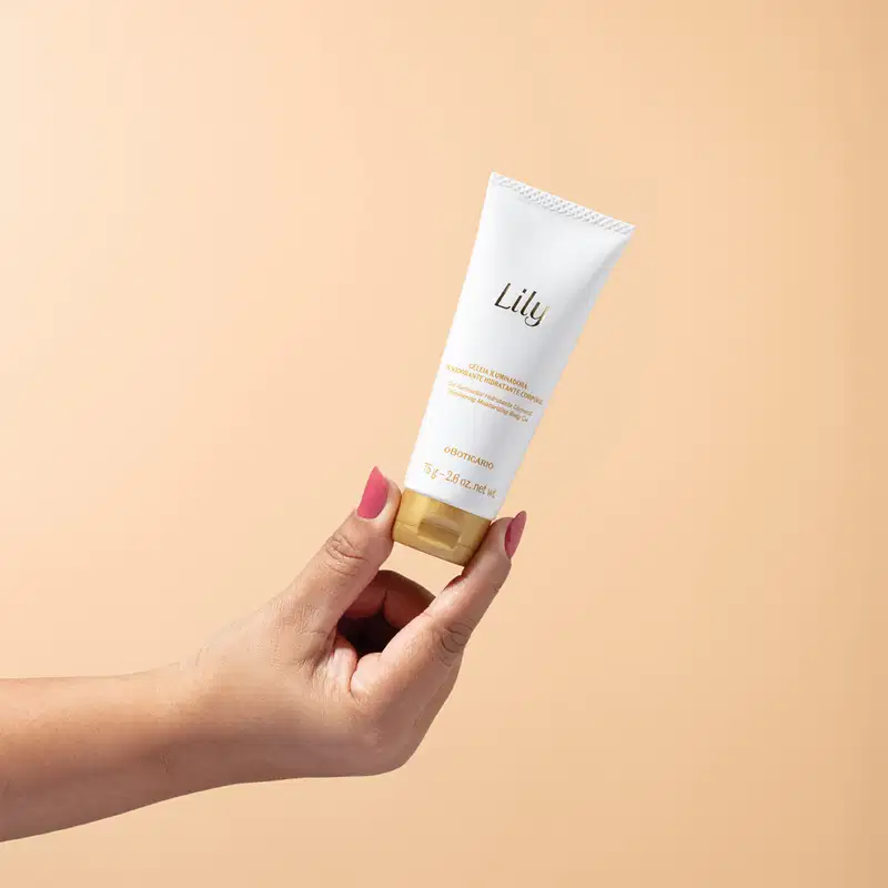 Mão feminina segurando um frasco branco com letras e tampa em dourado contendo 75g de geleia iluminadora hidratante corporal da marca Lily em fundo em tons de rosa.