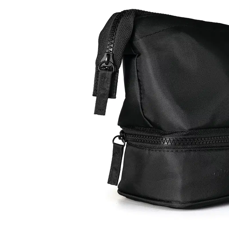 Close-up de Nécessaire masculina preta de nylon em formato triangular com foco na lateral que contém o puxador do zíper, com um puxador feito com pequena faixa retangular de nylon.