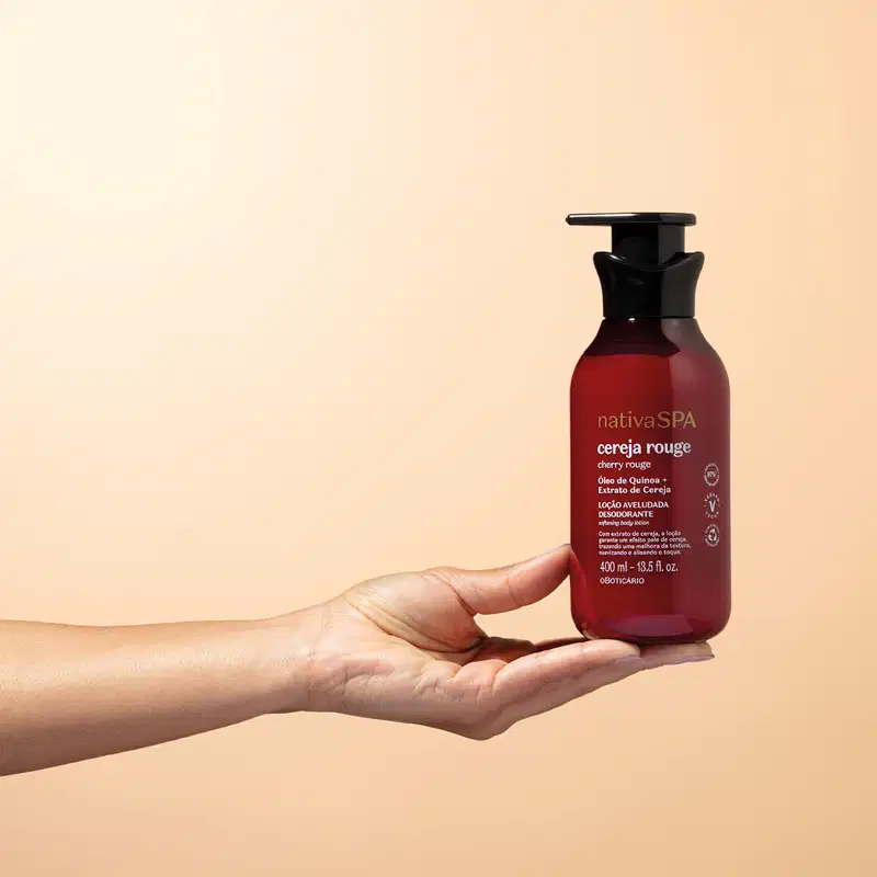 Mão feminina segurando um frasco vermelho escuro com tampa dosadora preta e letras douradas e brancas contendo loção aveludada corporal da marca Nativa SPA Cereja Rouge em fundo rosa claro.