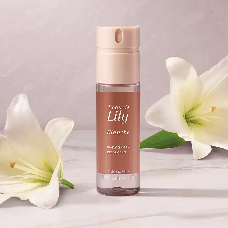 O frasco é de cor rosa claro, com um rótulo que exibe o nome da fragrância em destaque. O produto é apresentado em um recipiente com um aplicador spray. Ao lado do frasco, há flores de lírio brancas, que parecem complementar a imagem do produto.