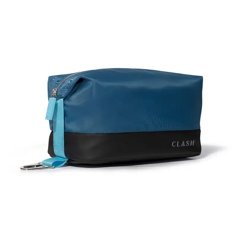 Bolsa de cosméticos azul-marinho com faixa preta na barra, com zíper e alça, exibindo a marca "CLASH" em letras grandes no canto inferior direito.  A vista lateral permite ver o detalhe em tecido azul mais claro do topo do zíper até a base do nécessaire.