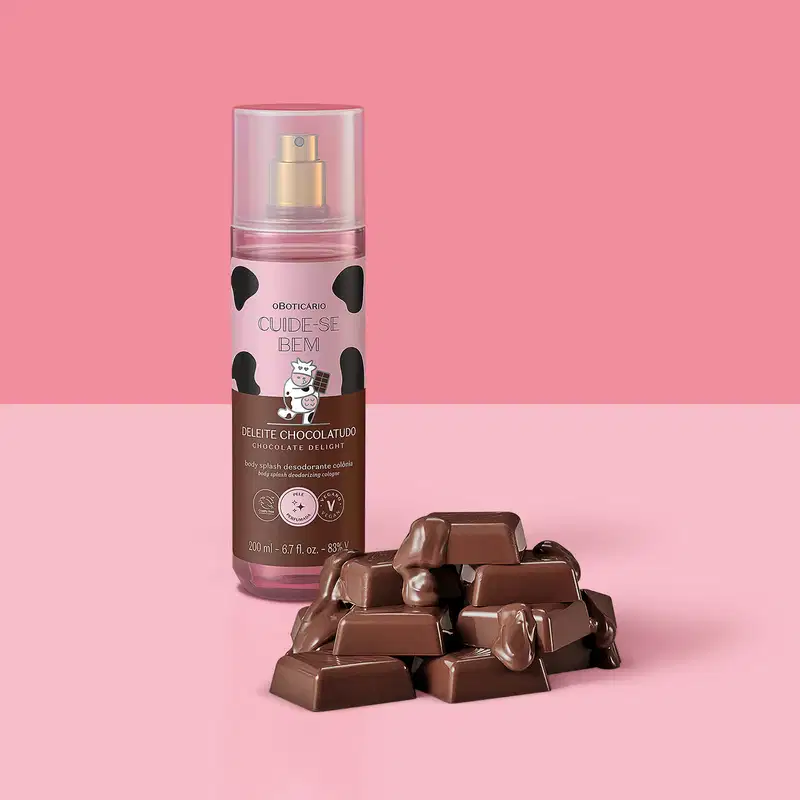 Frasco com rotulo rosa e marrom com vaca ilustrada de body splash Cuide-se Bem Deleite Chocolatudo com pedaços de chocolate ao leite espalhados ao lado do frasco, representando o aroma do produto em fundo rosa.