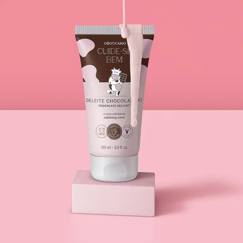 Frasco rosa e marrom com ilustração de uma vaquinha contendo creme esfoliante corporal Cuide-se Bem Deleite em uma plataforma branca com creme escorrendo sobre a embalagem, em fundo e superfície em tons de rosa.