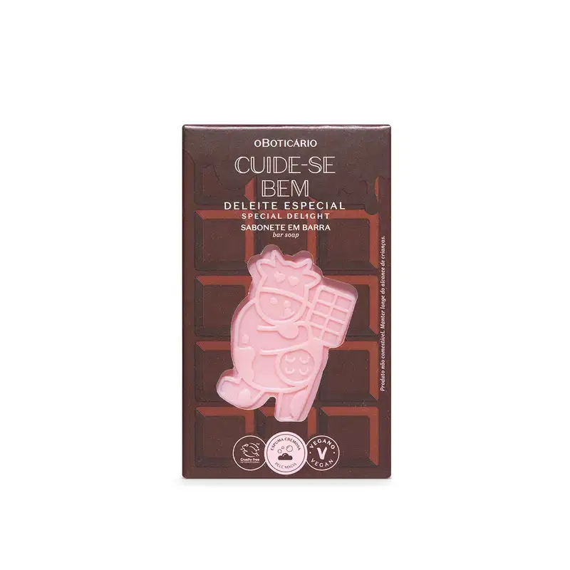 Sabonete em barra com vaca rosa estampada. Embalagem retangular marrom com texto "Cuide-se Bem Deleite Especial" e "Sabonete em Barra" em letras brancas. Selos indicando produto vegano e cruelty-free. Cores predominantes são marrom, rosa e branco.