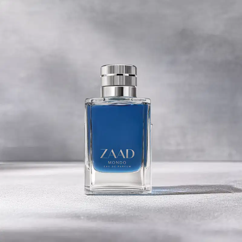 Frasco de vidro transparente de item de perfumaria da marca Zaad Mondo, contendo líquido na cor azul, tampa prateada e marca gravada no rótulo, também em prata sobre fundo cinza, com iluminação cênica, formando uma longa sombra do produto na superfície.