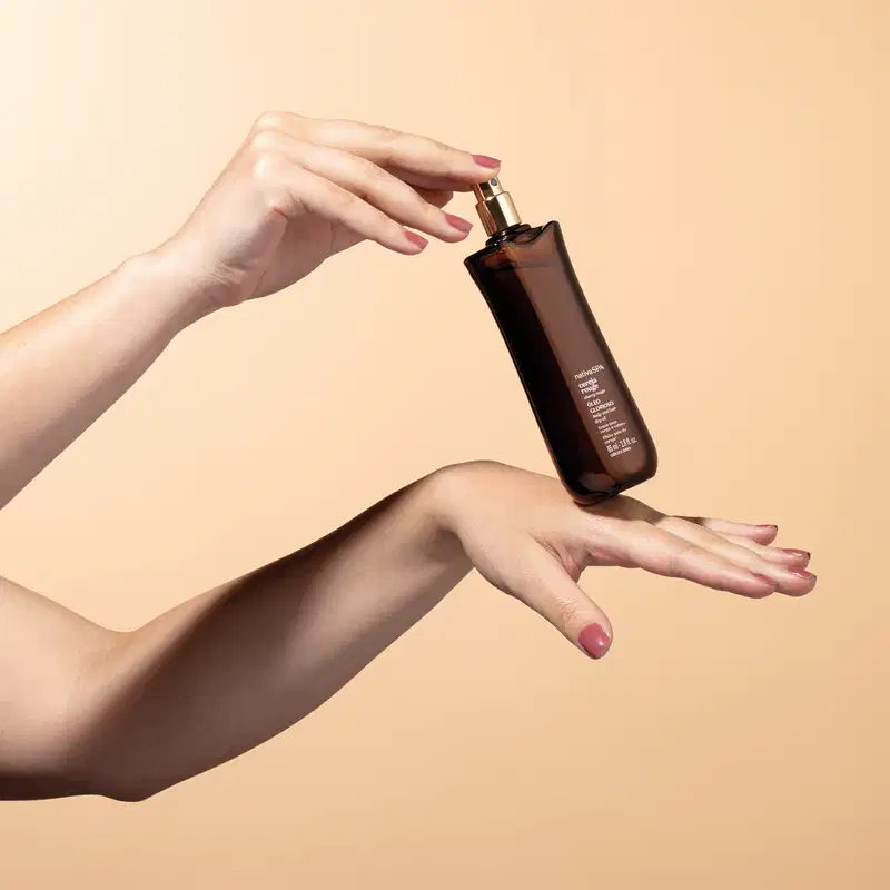 Mãos segurando um frasco de óleo glorioso para corpo e cabelos da marca Nativa SPA Cereja Rouge sobre fundo bege, com destaque para a embalagem escura e o gesto delicado.