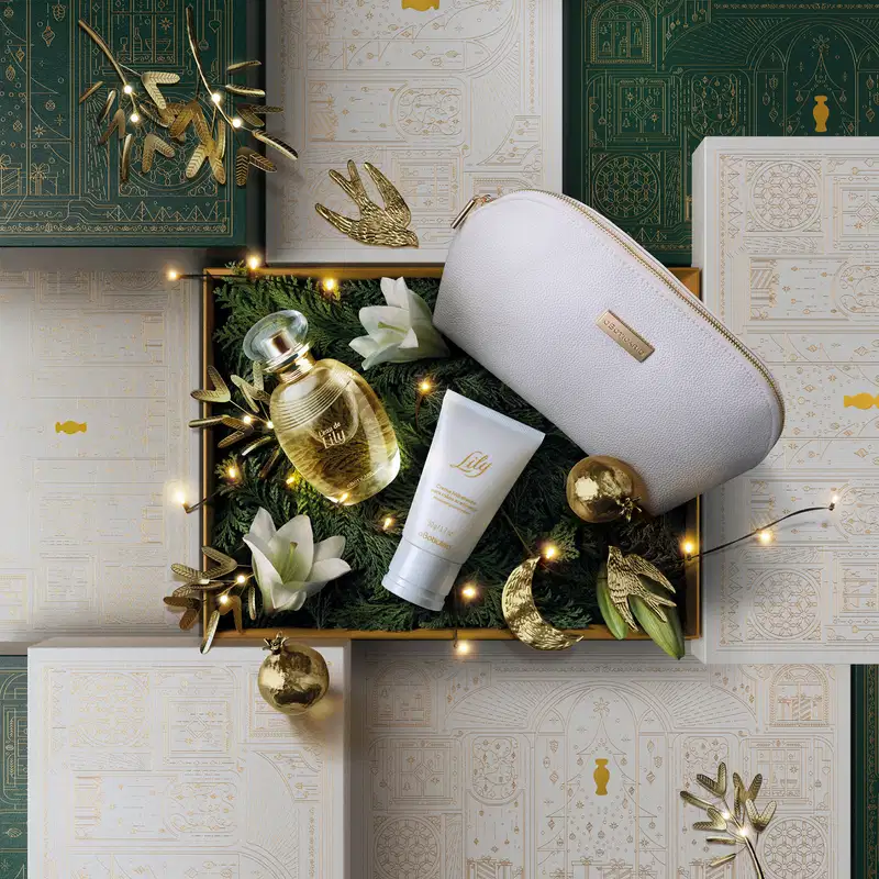 Caixa de presente bege com detalhes dourados, nécessaire azul, vidro com líquido amarelo com tampa dourada de L`eau De Lily e bisnaga branca com tampa cinza de creme de mãos acetinado da marca Lily em cenário natalino e muitas luzes.