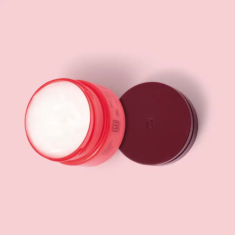 Pote aberto de Suflê Hidratante Desodorante Corporal da linha Egeo Choc High. A tampa vinho está sobre uma superfície rosa ao lado da embalagem, vistos de cima, o que permite ver o creme corporal branco dentro do pote rosa.