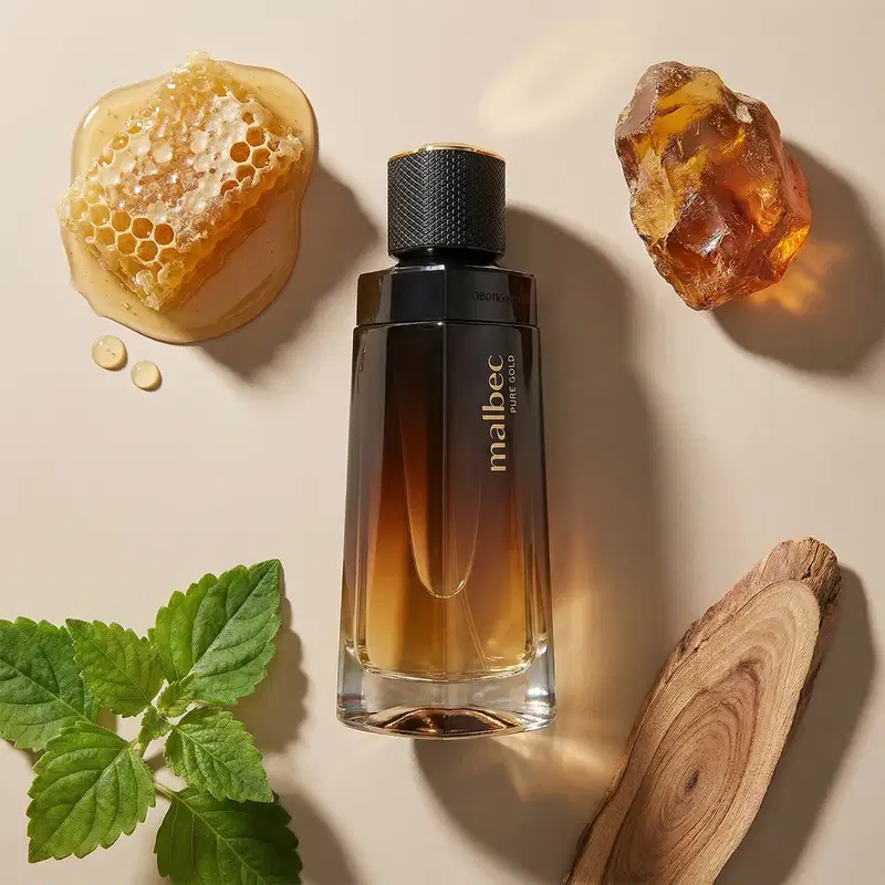 Imagem de um recipiente de desodorante colônia da marca Malbec Pure Gold em meio a elementos naturais como favo de mel, cristal de âmbar e folhas verdes, sobre superfície clara.