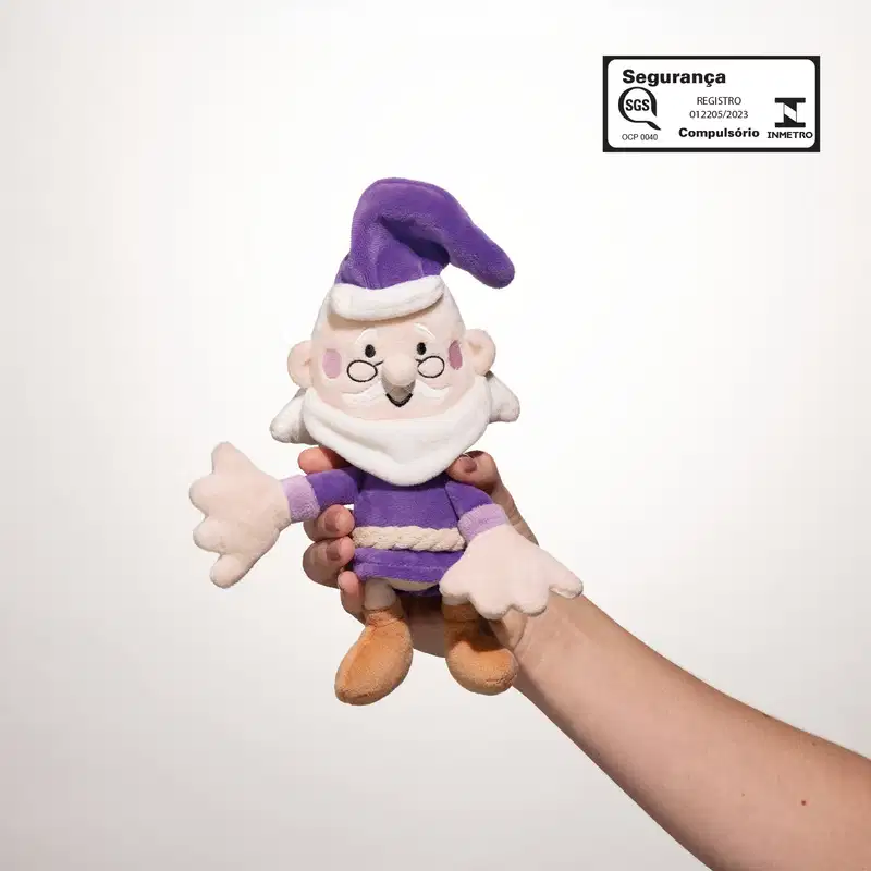 Mão segurando um boneco de tecido macio, personagem da marca Dr. Botica, um senhor de barba branca, óculos pretos, gorro e roupas roxas em um fundo claro.
