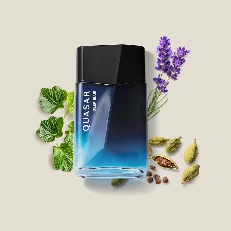 Frasco de desodorante colônia da marca Quasar Deep Blue em embalagem azul, rodeado por folhas verdes, flores roxas e especiarias em um ambiente claro e natural.