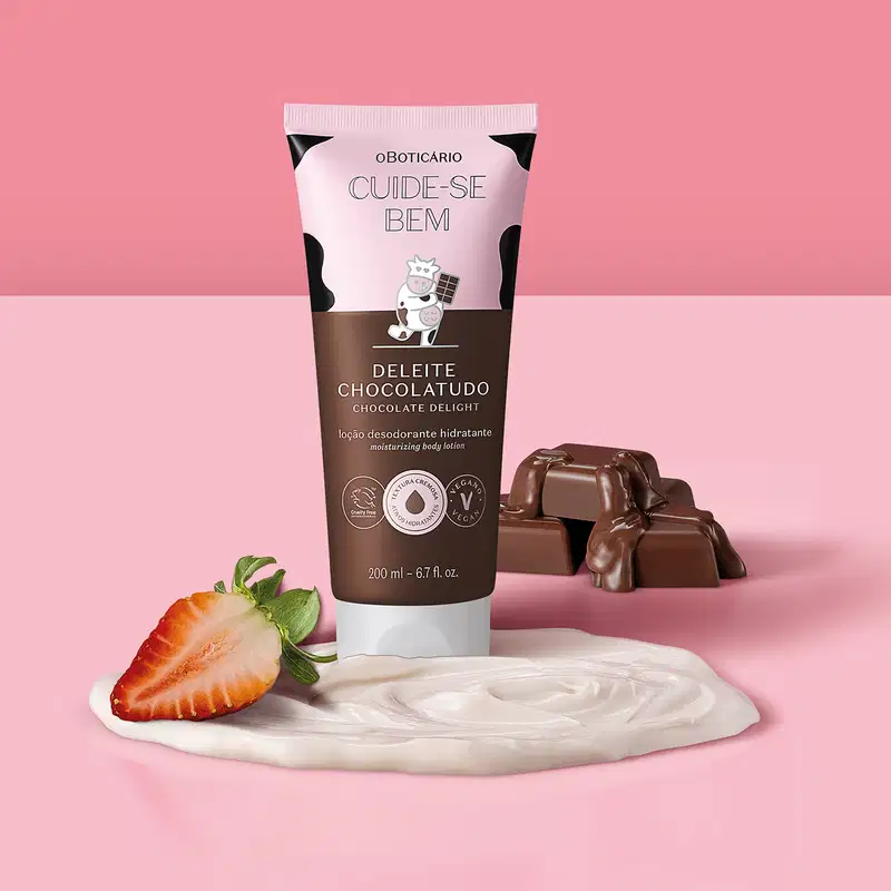 Embalagem de loção hidratante corporal Cuide-se Bem Deleite Chocolatudo ao lado de barras de chocolate, um morango cortado ao meio e um creme rosa claro, todos dispostos sobre um fundo rosa. 