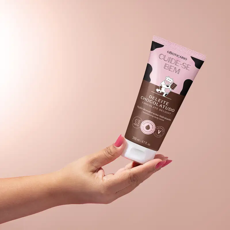 Frasco de loção hdratante corporal Cuide-se Bem Deleite Chocolatudo em rosa e marrom com vaca ilustrada sendo segurado por uma mão feminina em fundo lilás.