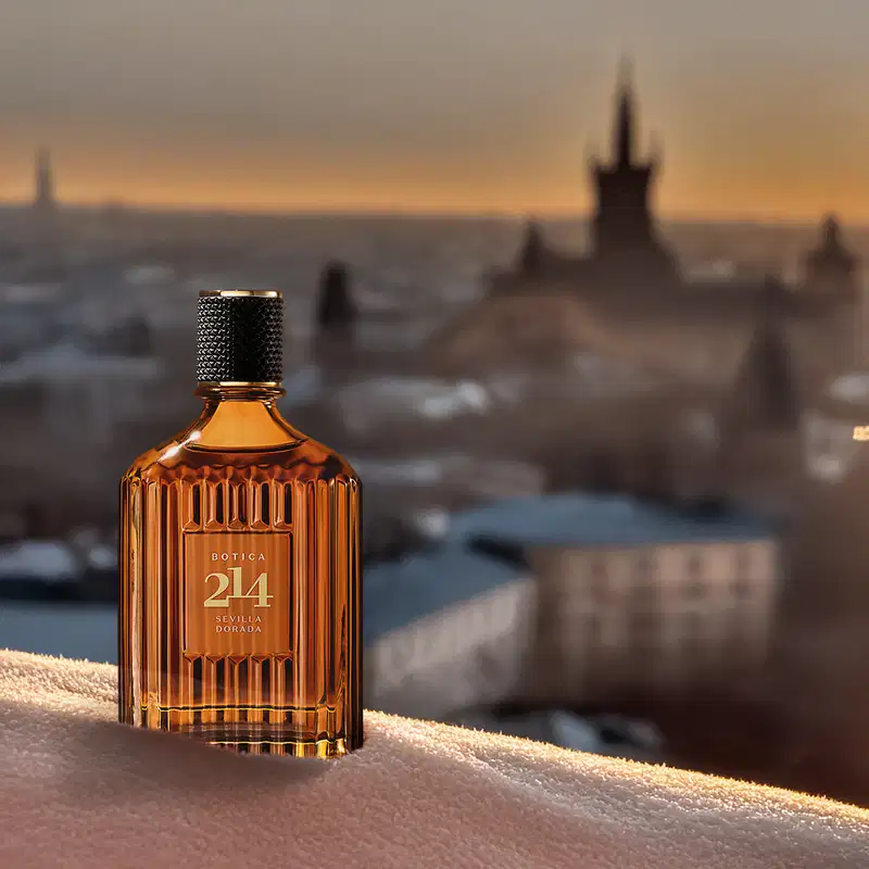 Frasco de Eau de Parfum Fougére Aromático Botica 214 Sevilla Dorada em primeiro plano, com paisagem de castelo e casas ao fundo em tons de laranja e azul durante o pôr do sol.
