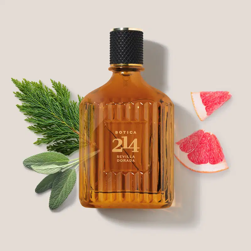 Frasco de Eau de Parfum Fougére Aromático Botica 214 Sevilla Dorada em fundo claro, com ramos de plantas verdes e fatias de toranja ao redor, criando um cenário natural e aromático.
