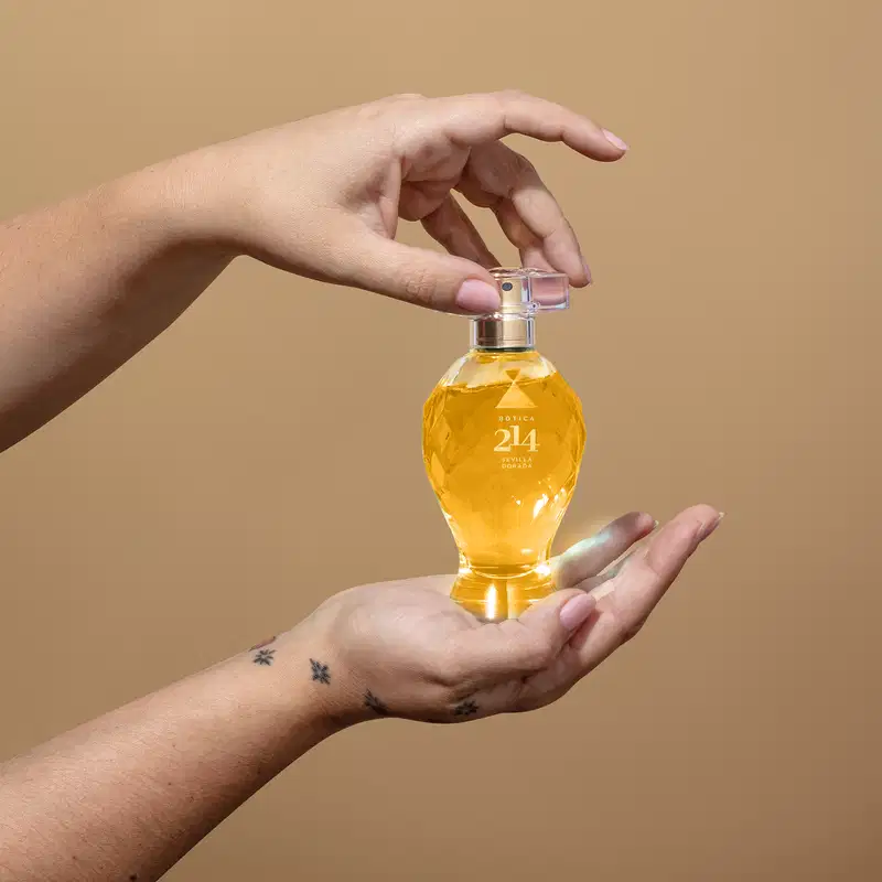 Frasco de Eau de Parfum Floral Frutal Botica 214 Sevilla Dorada em mãos femininas sobre fundo bege, com superfície lisa e iluminação suave.
