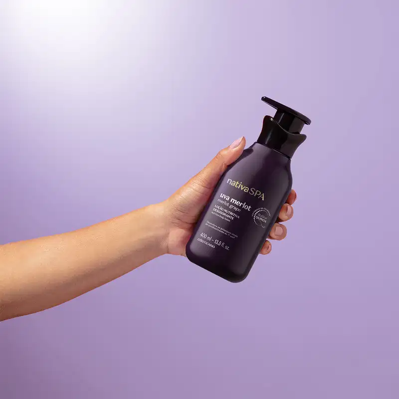 Mão feminina segurando um frasco roxo escuro e preto com letras douradas e brancas de loção hidratante corporal Nativa SPA Uva Merlot em fundo azul e branco.