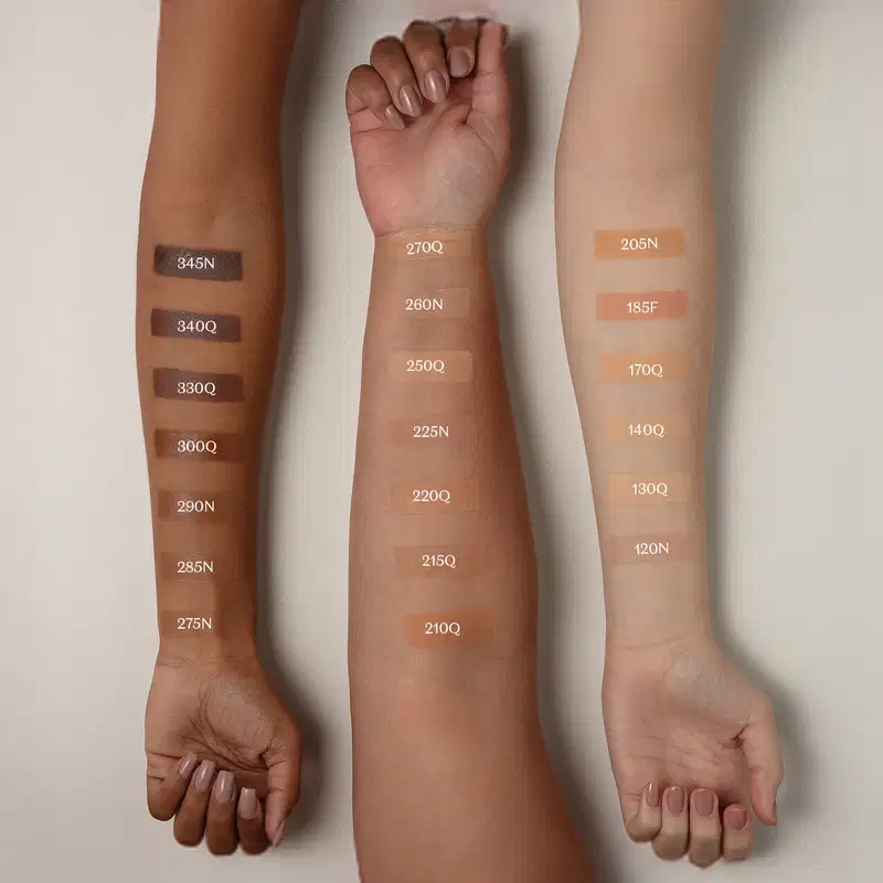 Imagem mostra três braços de cores diferentes estendidos sobre superfície branca sendo o braço mais a esquerda de pele negra, o do meio de pele parda e o da direita com pele branca, todos com diversos tons de base aplicados sobre eles e seus códigos.