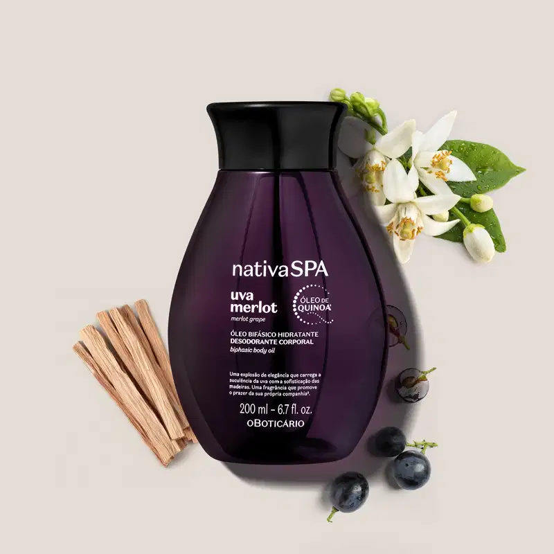 Frasco de óleo hidratante corporal da marca Nativa SPA Uva Merlot em embalagem roxa, com flores brancas e folhas verdes ao redor, sobre superfície clara com uvas e madeira ao redor.