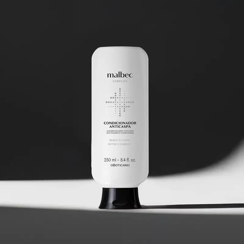 Imagem de frasco branco com tampa preta e rótulo com letras pretas da marca Malbec para cabelos, sobre superfície branca iluminada com luz cênica projetando uma sombra a direita do frasco sobre fundo preto.